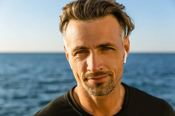 Portrait en gros plan d'un homme adulte souriant avec écouteurs sans fil sur le bord de la mer regardant une caméra — Photo de stock