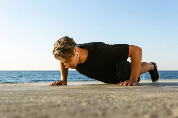 Sporty adult man with wireless earphones doing push ups on seashore — Stock Photo