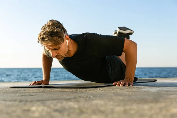 Fit adult man with wireless earphones doing push ups on knees on seashore — Stock Photo