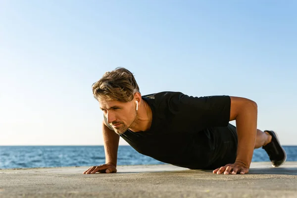 Handsome adult sportsman with wireless earphones doing push ups on seashore — Stock Photo