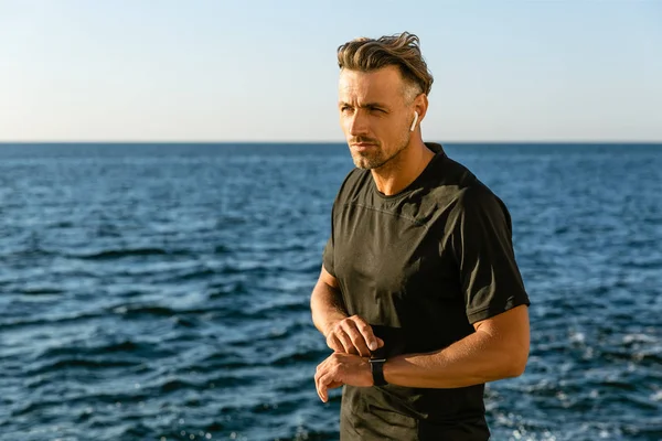 Hombre adulto guapo con auriculares inalámbricos y reloj inteligente en la orilla del mar - foto de stock