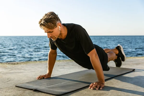 Sportivo adulto con auricolari wireless che fa flessioni in riva al mare — Foto stock