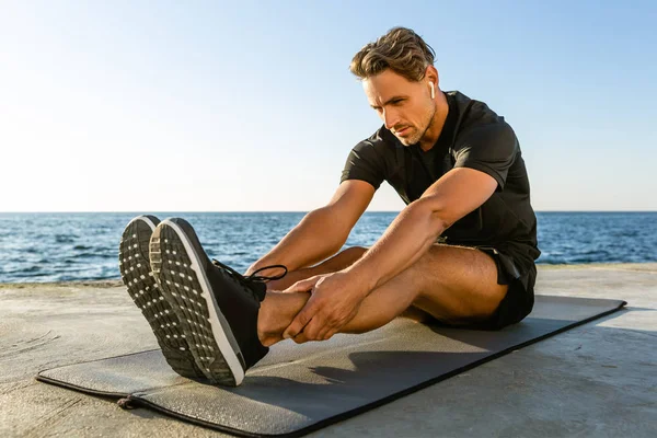 Adult sportsman with wireless earphones stretching on seashore — Stock Photo