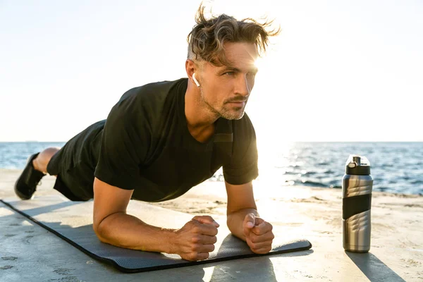 Apuesto deportista adulto con auriculares inalámbricos haciendo ejercicio tablón en la orilla del mar - foto de stock
