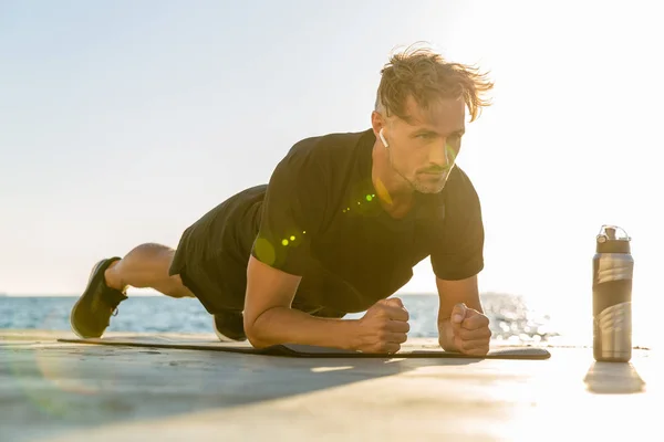 Fit adult man with wireless earphones doing plank exercise on seashore — Stock Photo