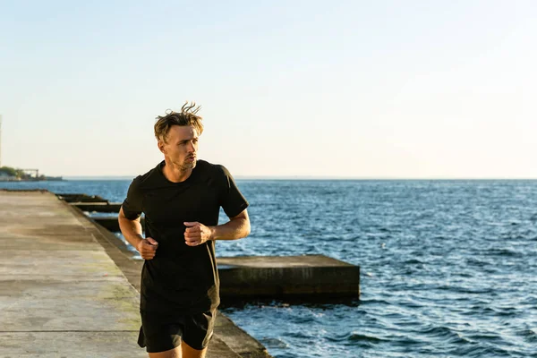 Atlético adulto hombre en negro sportswear jogging en la orilla del mar en la mañana — Stock Photo
