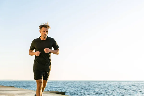 Atlético joger adulto corriendo en la orilla del mar en la mañana - foto de stock