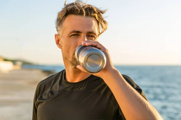 Hombre adulto guapo bebiendo agua de la botella de la aptitud en la orilla del mar delante de la salida del sol después del entrenamiento - foto de stock