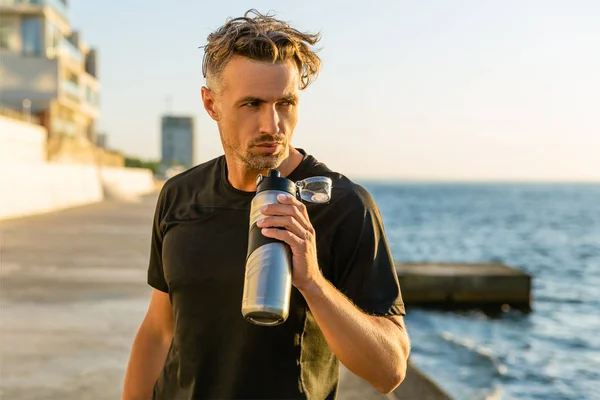 Apuesto deportista adulto con pelo gris sosteniendo botella de fitness en la orilla del mar delante de la salida del sol mirando hacia otro lado - foto de stock