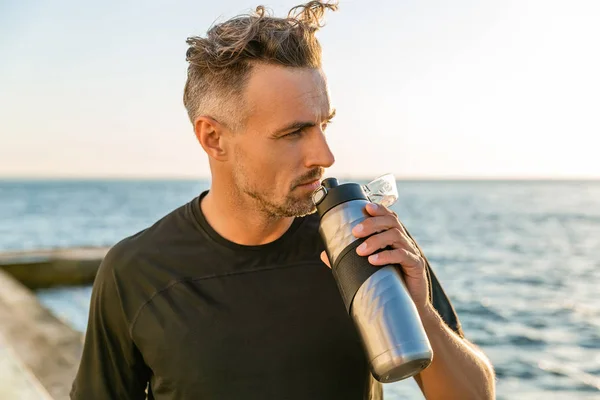 Deportista adulto con pelo gris sosteniendo botella de fitness en la orilla del mar frente a la salida del sol mirando hacia otro lado - foto de stock