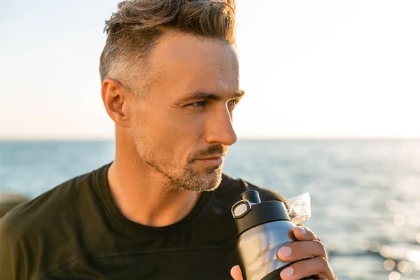 Close-up portrait of handsome adult man drinking water from fitness bottle on seashore in front of sunrise — Stock Photo