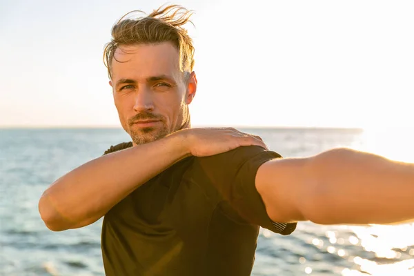 Athletic adult man stretching arm before training on seashore — Stock Photo