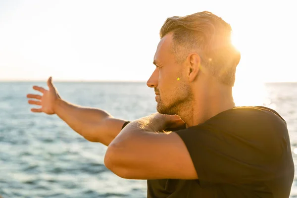Beau sportif adulte étirant le bras avant de s'entraîner au bord de la mer devant le lever du soleil — Photo de stock