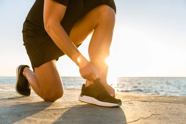 Cropped shot of sportsman lacing up sneakers on seashore in front of sunrise — Stock Photo