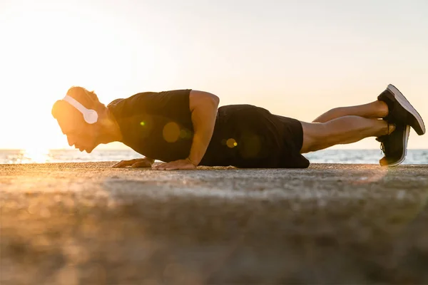 Seitenansicht eines gut aussehenden erwachsenen Sportlers mit Kopfhörern, der Liegestütze am Strand macht — Stockfoto