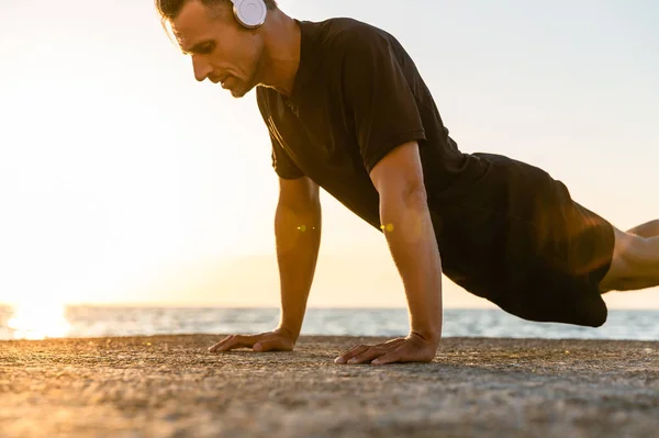 Athletic adult man in headphones doing push ups on seashore — Stock Photo