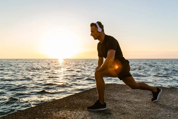 Atlético hombre adulto en auriculares haciendo sentadillas de una pierna durante el entrenamiento en la orilla del mar - foto de stock