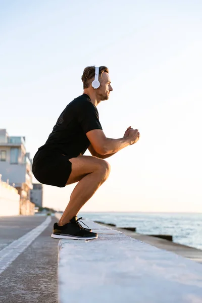 Vue latérale de l'homme adulte athlétique dans les écouteurs debout squats sur parapet sur le bord de la mer — Photo de stock