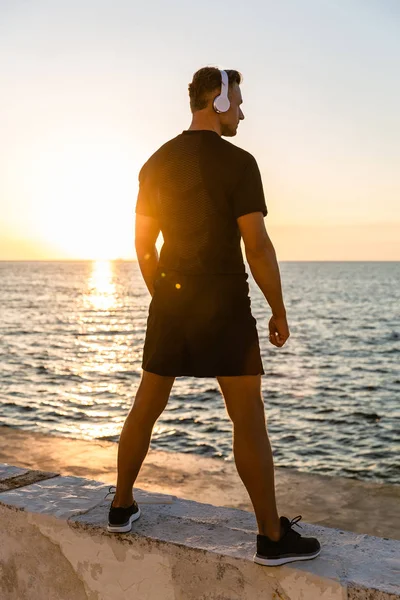 Rear view of athletic adult man in headphones standing on seashore in front of sunrise — Stock Photo