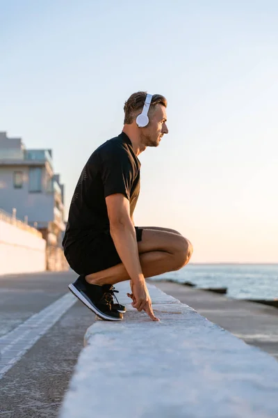 Vue latérale du beau sportif adulte dans des écouteurs debout squats sur parapet sur le bord de la mer — Photo de stock