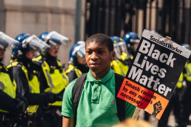 London / UK - 06/13/2020: Black Lives Matter protest during lockdown coronavirus pandemic. Young man with BLM banner posing in front of Police officers clipart