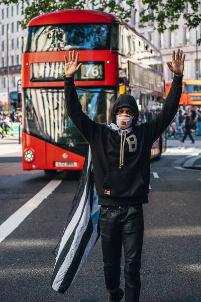 Londra / İngiltere - 06 / 13 / 2020: Siyahi Yaşamı Önemlidir protestosu koronavirüs salgını sırasında. Amerikan bayrağı pelerinli bir adam otobüsün önünde elleri havada yürüyor.