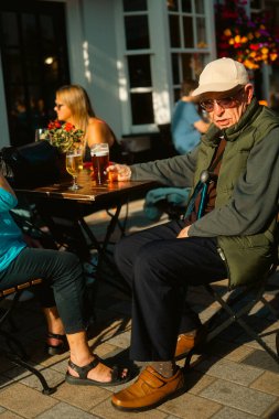 Twickenham, Londra, Birleşik Krallık 2025.09.06 güneş gözlüklü ve şapkalı yaşlı bir adam açık bir bar masasında oturmuş, öğleden sonra güneşinin tadını çıkarırken etrafındaki insanlarla birlikte.. 
