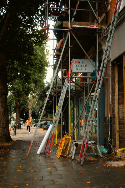 Chiswick, London, UK 2025.08.26 Работник на лесах возле филиала Barnard Marcus с лестницами и инструментами, видимыми на тротуаре под осенними деревьями. 