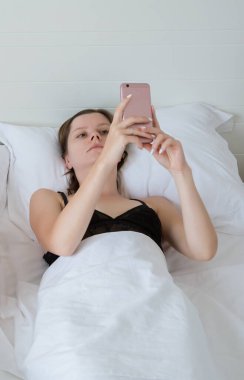 Yatakta cep telefonlu bir kadın son haberleri izliyor. Genç yetişkin bir kız otelde beyaz çarşaflarda uzanıyor ve telefonla sosyal medya ağını izliyor..