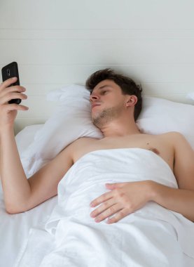Yatakta cep telefonlu bir adam en son yenisini izliyor. Genç adam otelde beyaz çarşaflarda uzanıyor ve telefonla sosyal medya ağını izliyor..
