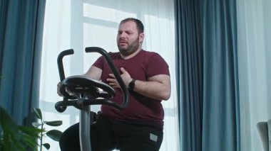 Aşırı kilolu bir adam evde bisiklet eğitimi alıyor ve hasta hissediyor, kardiyo egzersizi yapıyor. Yüksek kalite 4k görüntü
