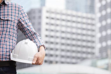 Mühendislik erkek inşaat işçisi standı ön görünümü emniyet beyaz kask tutarak stand ve iş operasyonu güvenliği için yansıtıcı giyim giymek. Bina arka plan açık.