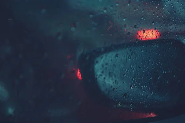 Car window with rain drops window on glass rearview mirror with road light bokeh glass background blurred. City life in night in rainy season night storm raining car driving concept. soft focus.