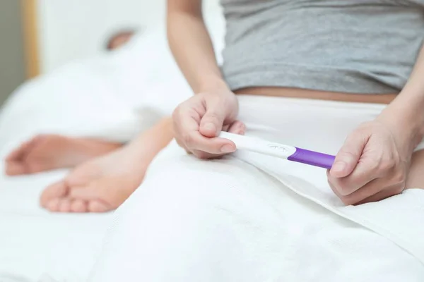 Young woman looking show in hand at checking pregnancy test kit. She\'s is checking her pregnancy exam sitting on the bed with couple lovers in bedroom. Premature is pregnant before age.