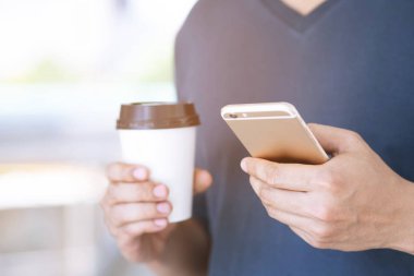 Cep telefonu kullanan genç erkek, mola eli tutan kağıt fincan ıstıranın sabah ları parkta sıcak kahve içerken cep telefonundan mesaj izlerken duruyor.