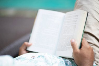 Rahat giysiler içinde genç adam geri görünümü açılış ve bir kitap okuma ve yüzme havuzu nun yanında kanepede yatarken rahatlatıcı.