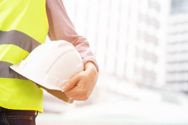 Mühendislik erkek inşaat işçisi standı ön görünümü emniyet beyaz kask tutarak stand ve iş operasyonu güvenliği için yansıtıcı giyim giymek. Bina arka plan açık.