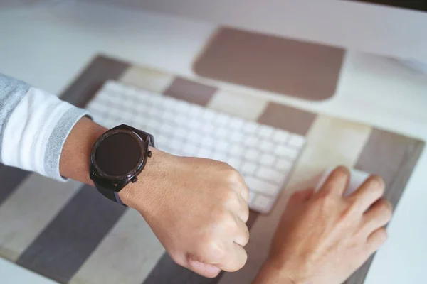 young man looking at his smart watch on his hand, watching the time. while working with mouse and keyboard computer laptop on wooden desk in office modern lifestyle.