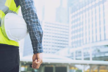 Mühendislik erkek inşaat işçisi standı nın arka görünümüne yakın iş çalışmasının güvenliği için güvenlik beyaz kask tutarak. bina arka plan açık.