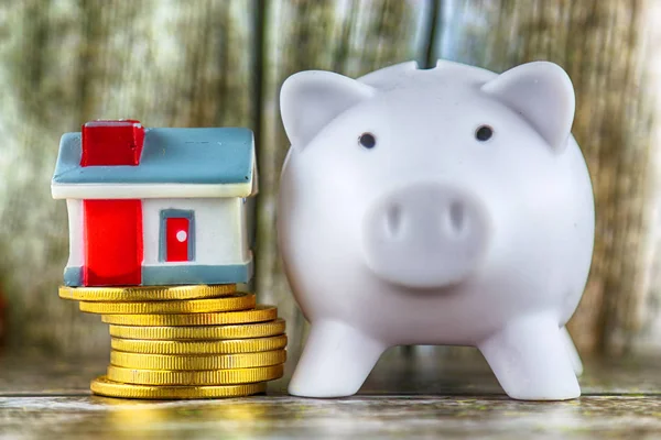 Piggy bank, miniature house and golden coins on wooden background. Savings in the household.