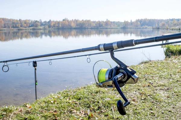 Carp rods are installed on the stands on the shore against the flat surface of the lake with a coil in the foreground. Background