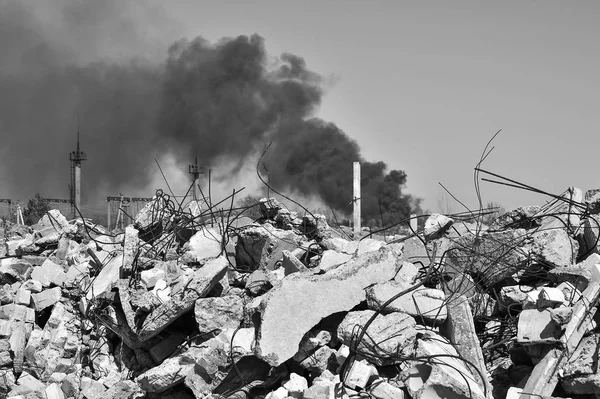 Ein Haufen Betonschutt mit auskragenden Bewehrungsstäben vor dem Hintergrund dicken schwarzen Rauchs am Himmel. Hintergrund. Schwarz-Weiß-Bild — Stockfoto