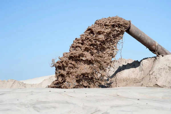 The flow of brown liquid from the pipe on the background of a pile of sand and blue clear sky. — Stock Photo, Image