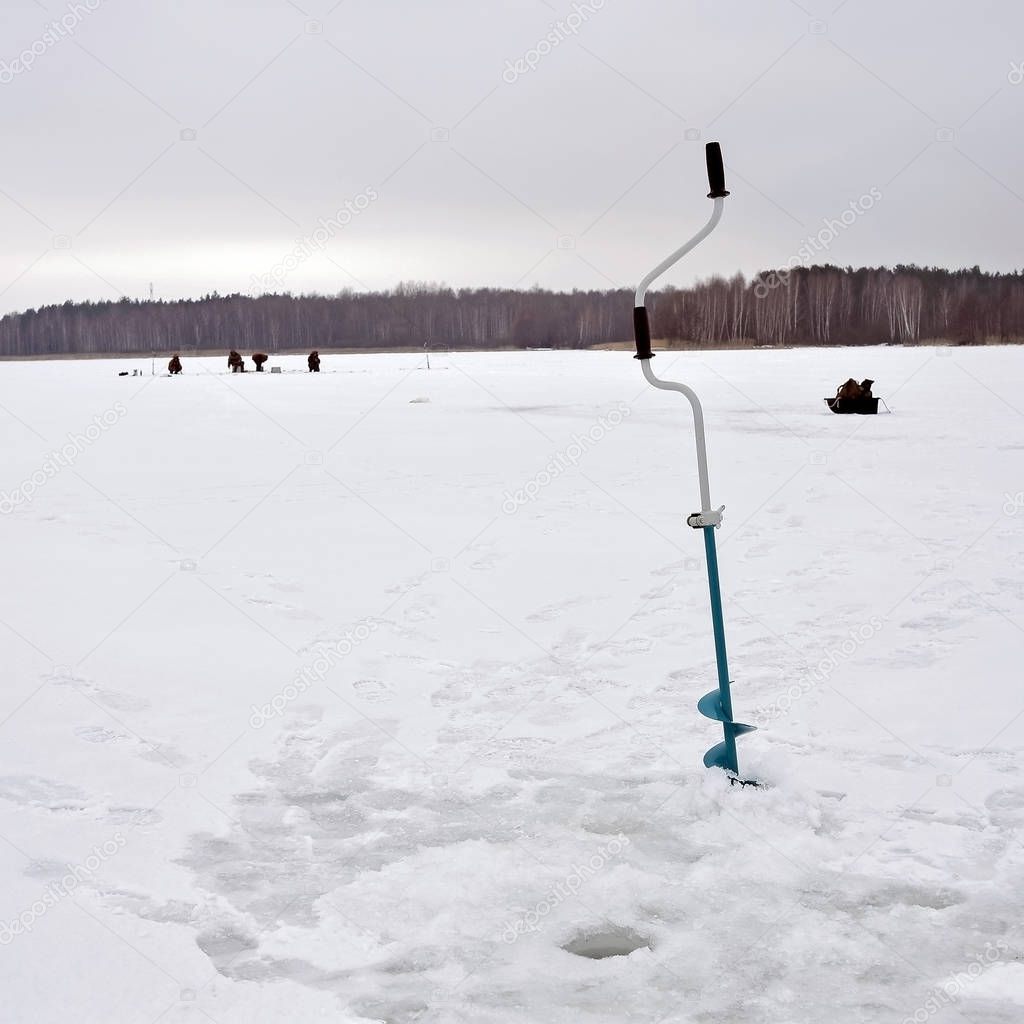 Ice drill hammered into the ice with a drilled hole on the background of a group of fishermen on a frozen lake