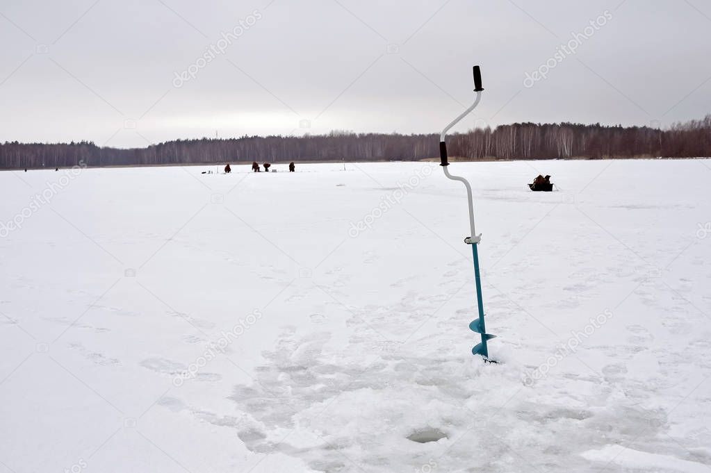 Ice drill hammered into the ice with a drilled hole on the background of a group of fishermen on a frozen lake