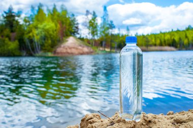Kapalı kapaklı mavi bir plastik şişe, kumlu bir sahilde vahşi bir gölün arka planında duruyor. Arkaplan.