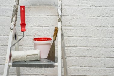 Beyaz bir duvarın arkasındaki metal bir merdiven üzerinde tuğla tezgahın altında bir spatula, kova ve dokuma rulosu..