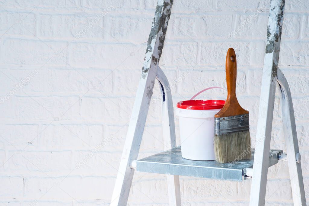 A paintbrush and bucket with a red lid stand on a metal stepladder against a white wall decorated with bricks. Background.