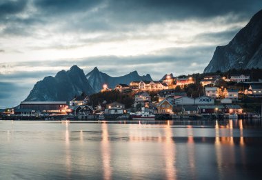 Norwegian fishing village Reine at the Lofoten Islands in Norway. Dramatic sunset clouds moving over steep mountain peaks in dusk. Reflection of street lights in atlantic Ocean. clipart
