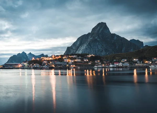 Norska fiskebyn Reine på ögruppen Lofoten i Norge. Dramatisk solnedgång moln flyttar över branta bergstoppar i skymning. Reflektion av gatubelysningen i Atlanten. — Stockfoto